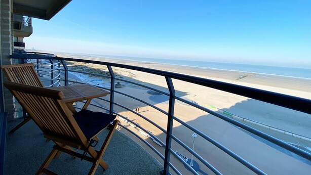 A balcony with a wooden chair and table, overlooking a beach with a clear sky. - Apartment in Nieuwpoort Near the Beach (Nieuwpoort)