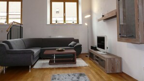 A modern living room with a grey sectional sofa, a wooden coffee table, a flat-screen TV on a wooden stand, and a large arched window with blinds. - Kampenwand Loft Modern Retreat (Aschau im Chiemgau)
