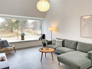 A modern living room with a sofa, a round wooden coffee table, a floor lamp, and a view of the outdoors through large windows.