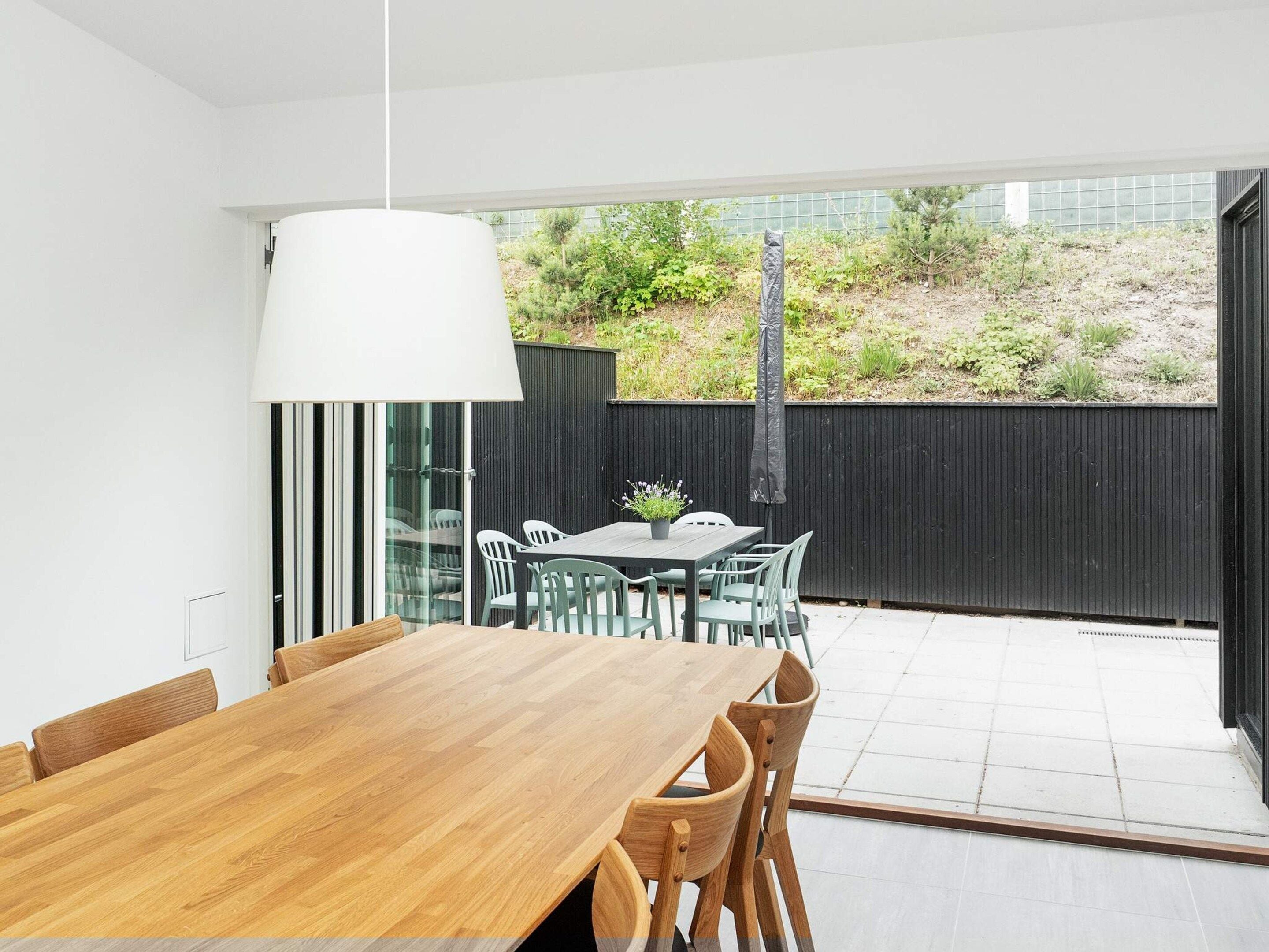 A modern dining area with a wooden table and chairs, a hanging light fixture, and a view of a garden through a glass door.