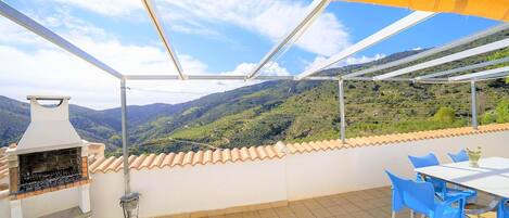 An outdoor patio with a barbecue grill, a table with chairs, and a view of a hilly landscape.