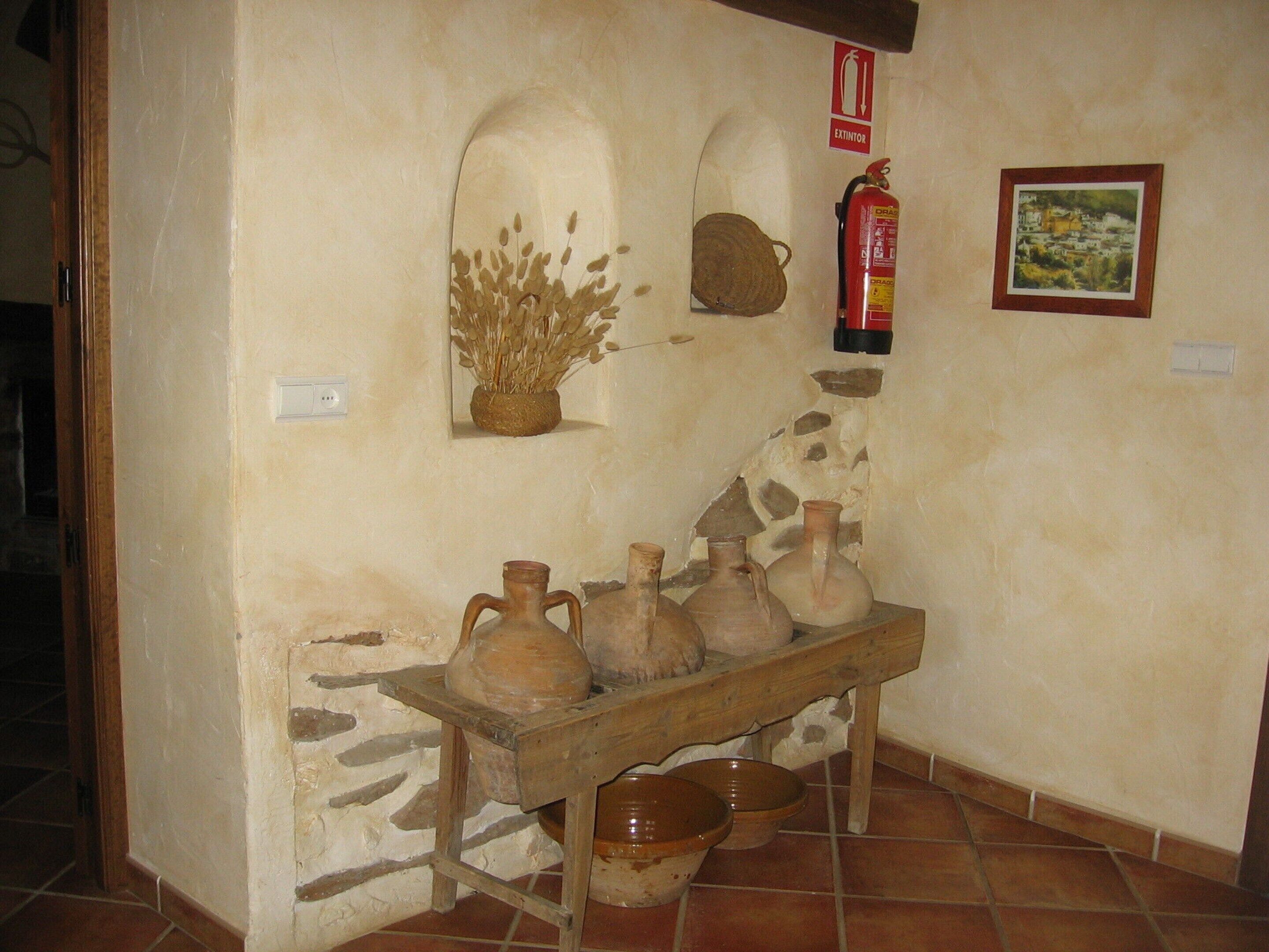A rustic interior with a stone wall, a wooden table holding pottery, a fire extinguisher, and a framed picture.