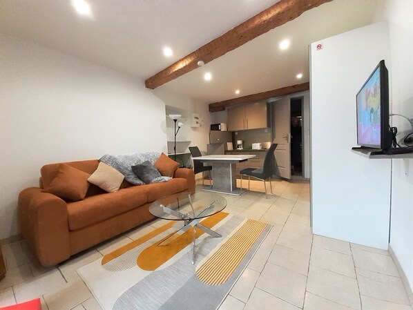 Uma sala de estar moderna com um sofá, uma mesa de centro de vidro e uma área de cozinha ao fundo.