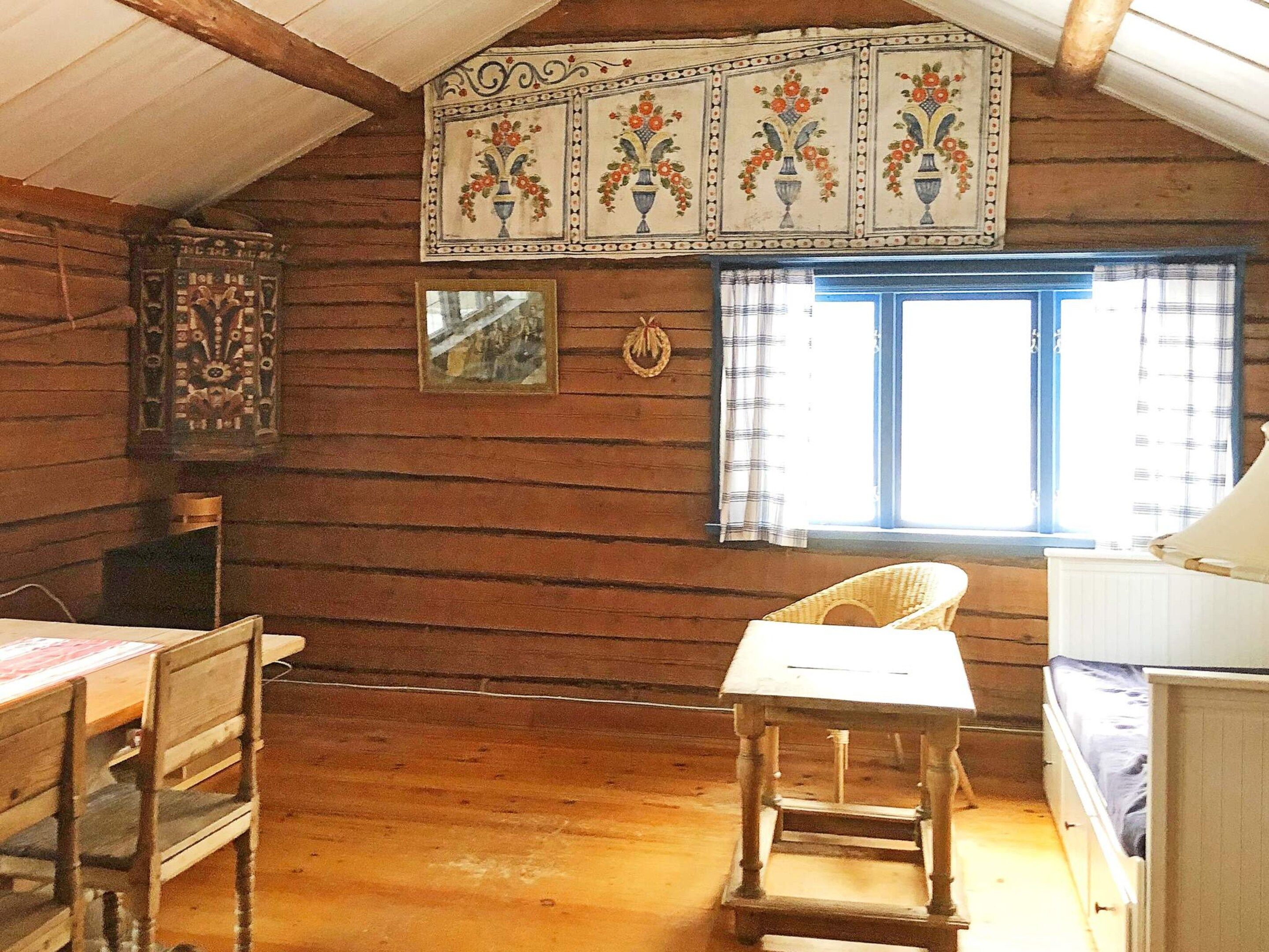 A rustic cabin interior with wooden walls, a dining table, a chair, a window with curtains, and a decorative wall hanging.