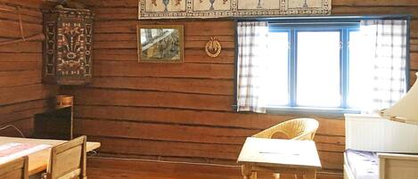 A rustic cabin interior with wooden walls, a dining table, a chair, a window with curtains, and a decorative wall hanging.