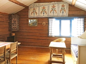 A rustic cabin interior with wooden walls, a dining table, a chair, a window with curtains, and a decorative wall hanging.