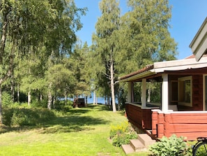 A wooden cabin with a porch, surrounded by lush greenery and trees, overlooking a body of water.