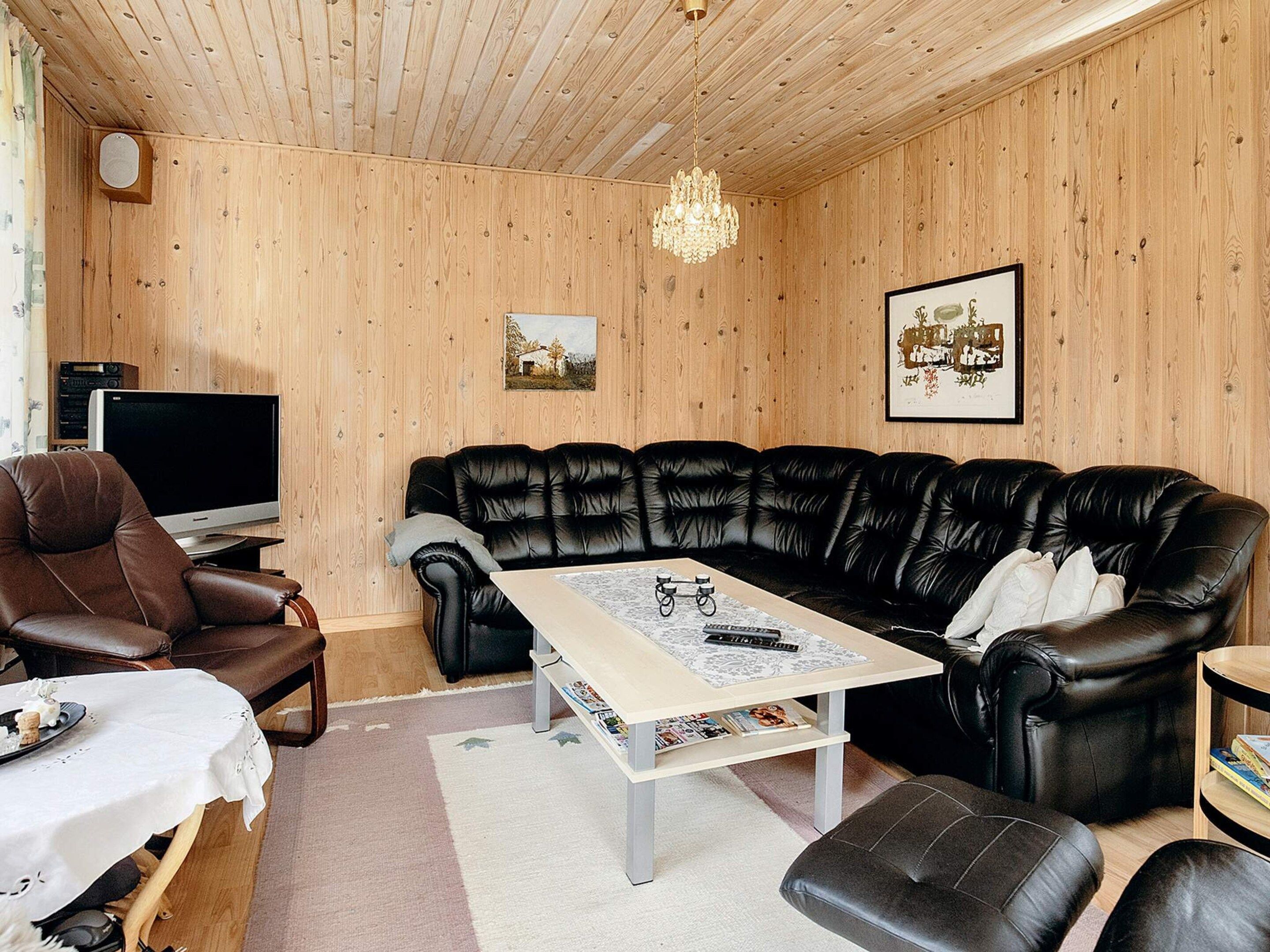 A living room with a black leather sofa, a brown leather armchair, a wooden ceiling, a chandelier, and a framed picture on the wall.