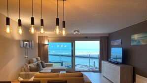 Una sala moderna con un sofá grande, una mesa de comedor y un televisor. Se ve el océano a través de amplios ventanales.