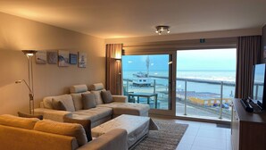 Una sala de estar moderna con un sofá, un televisor de pantalla plana y vista al mar a través de ventanas amplias.