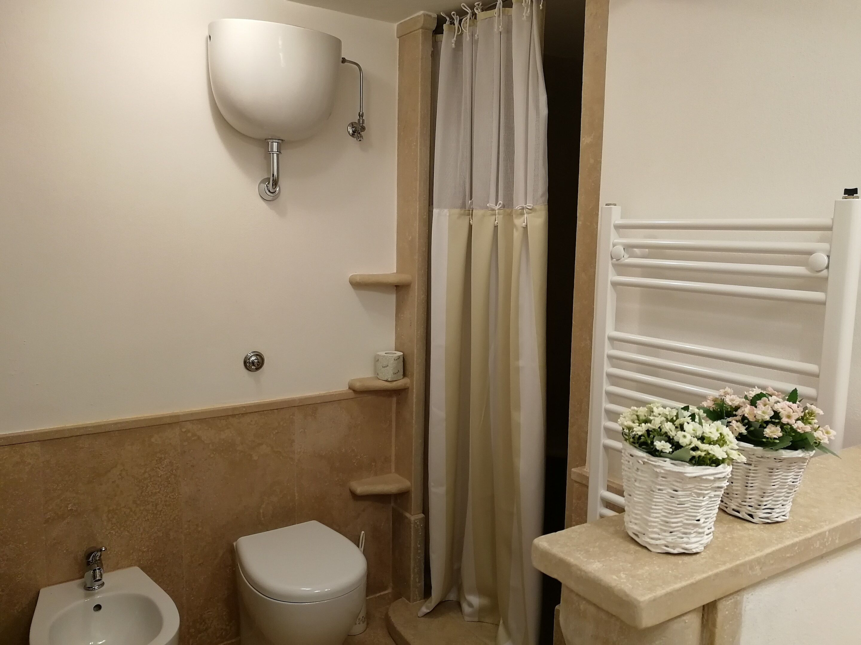 Une salle de bain avec un lavabo, des toilettes et un bidet, tous de couleur blanche. On y trouve deux paniers en osier remplis de fleurs, posés sur le comptoir.