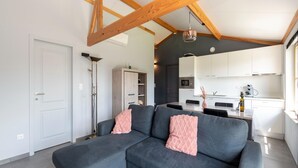 A modern living room with a grey sectional sofa, a white door, and a kitchen in the background. - Holiday Home in Koksijde With Private Garden (Koksijde)