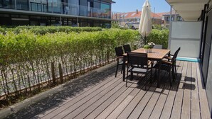 A wooden deck with a table and chairs, surrounded by a hedge and a modern building with balconies. - Apartment Nieuwpoort Near Beach (Nieuwpoort)
