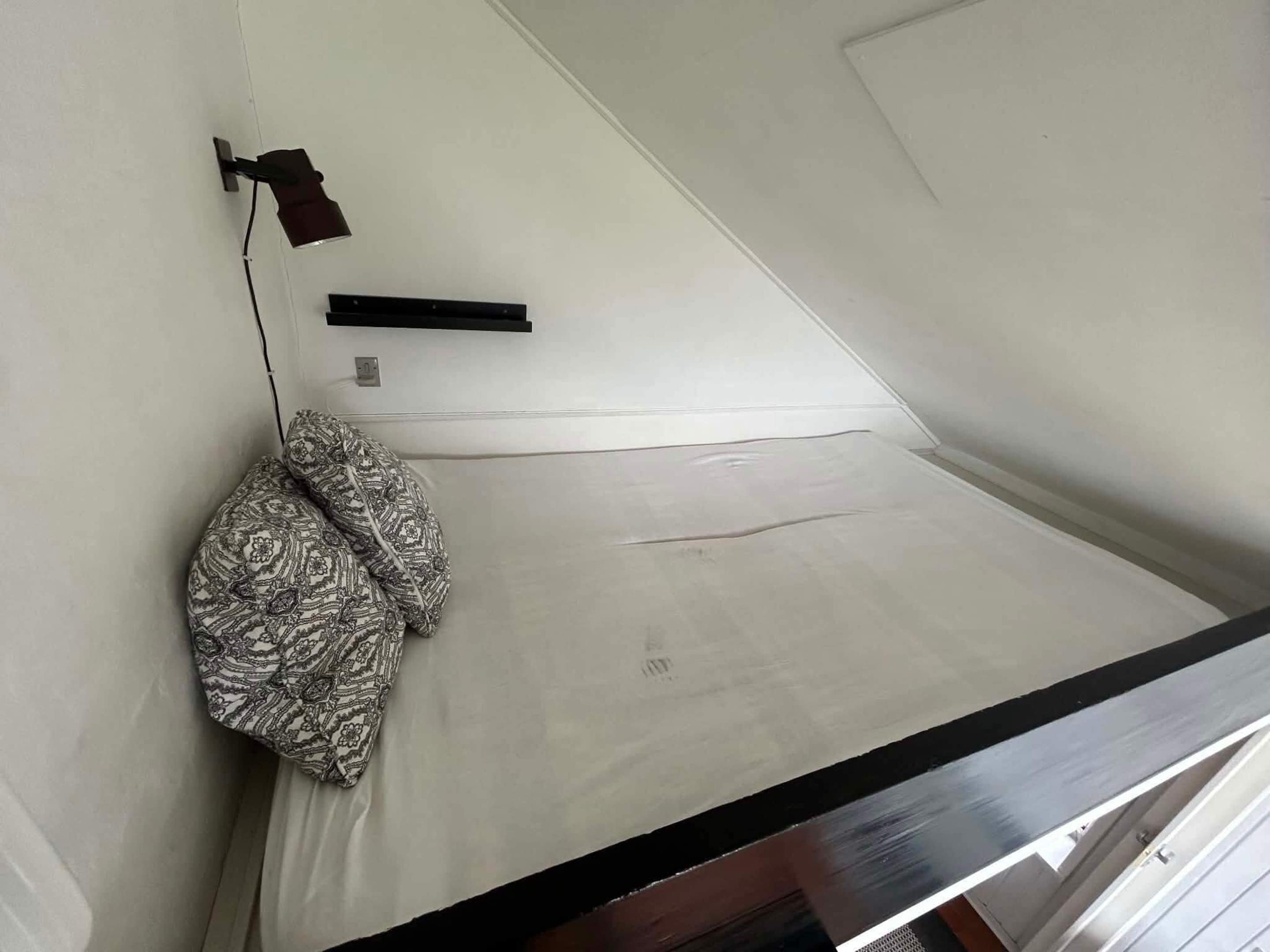 A room with a bed, a pillow, a wall-mounted lamp, and a small shelf.