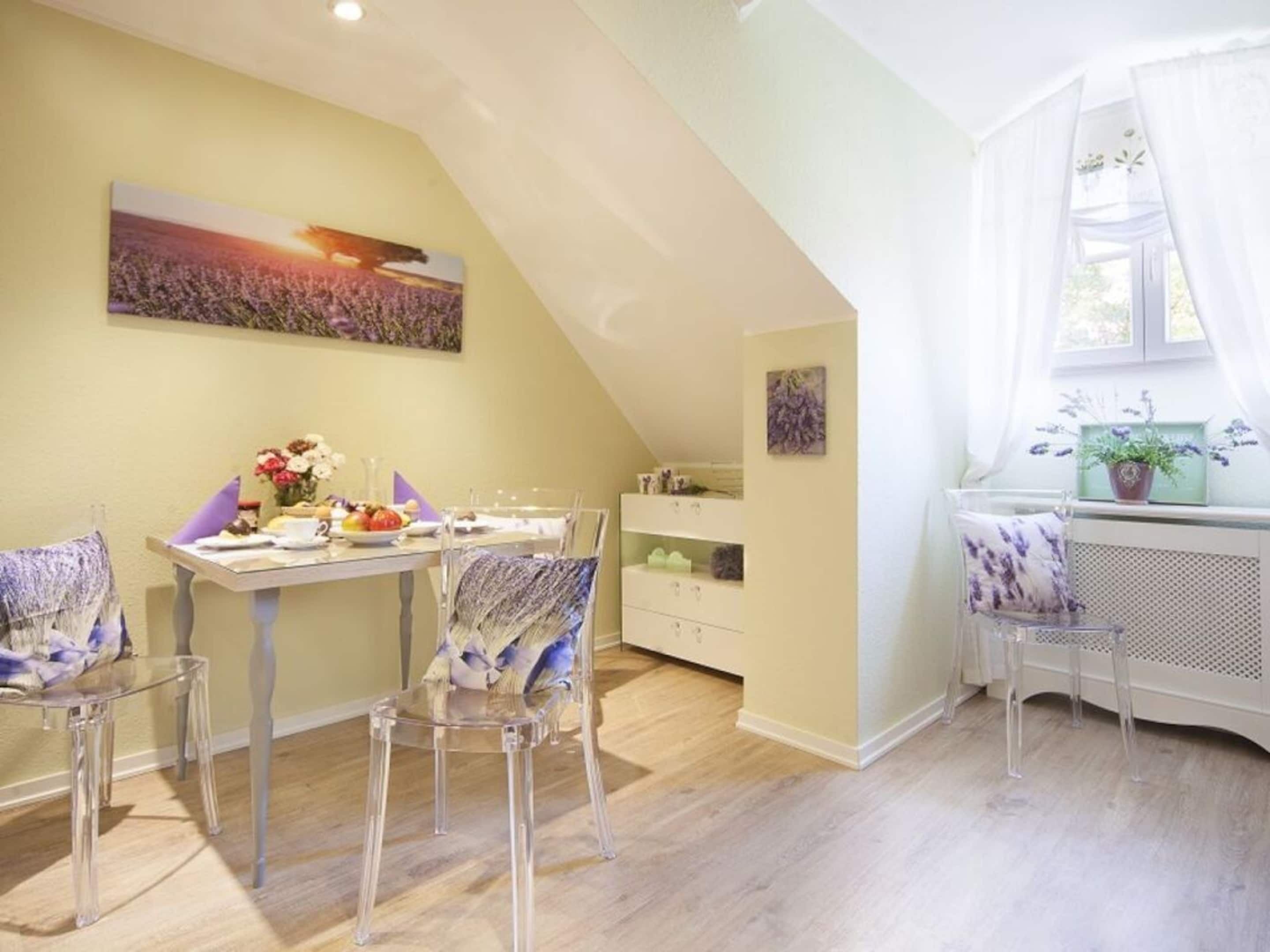 Un coin repas avec une table dressée pour deux personnes, des chaises transparentes, un tableau accroché au mur et une fenêtre avec des rideaux.