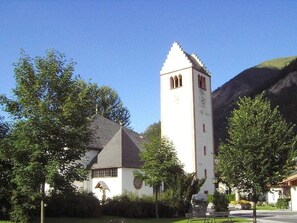 A church with a prominent bell tower and a clock, surrounded by trees and a mountain in the background. - Present Apartment in Fusch (Fusch an der Grossglocknerstrasse)