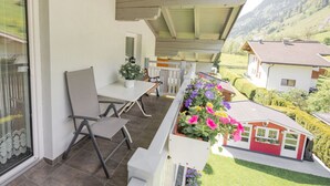 A balcony with a table and chairs, a small garden with flowers, and a view of a red house and green hills in the background. - Present Apartment in Fusch (Fusch an der Grossglocknerstrasse)
