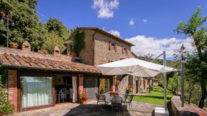A stone building with a tiled roof, a patio with a table and chairs, and a large umbrella. - Villa in Cortona With Pool & Hill Views (Cortona)