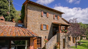 A stone house with a tiled roof, a glass door, and a small garden with a tree. - Villa in Cortona With Pool & Hill Views (Cortona)