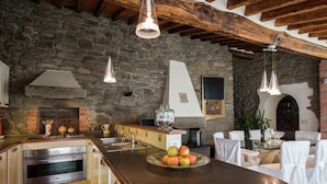 A kitchen with a stone wall, wooden beams, and a large island with fruit. - Villa in Cortona With Pool & Hill Views (Cortona)