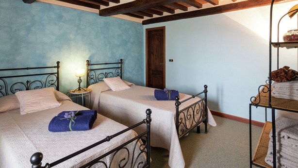 A bedroom with two beds, wooden beams on the ceiling, a nightstand with a lamp, and a shelf with towels. - Villa in Cortona With Pool & Hill Views (Cortona)