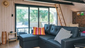 Ein gemütliches Wohnzimmer mit einem dunkelgrauen Ecksofa, einem kleinen Beistelltisch und einem Korbsessel. Ein großes Fenster bietet einen Blick ins Freie.