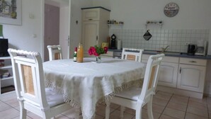 Un comedor con una mesa redonda cubierta por un mantel blanco, rodeada por cuatro sillas blancas con respaldo trenzado. En la mesa hay una botella de jugo de naranja y un florero con flores rosas.