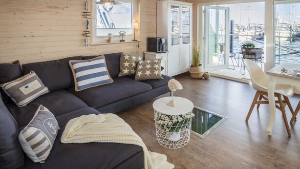 A modern living room with a dark sofa, a round coffee table, and a view of a marina through large windows. - Houseboat Grey Seal Comfortable Vacation Home (Heiligenhafen)