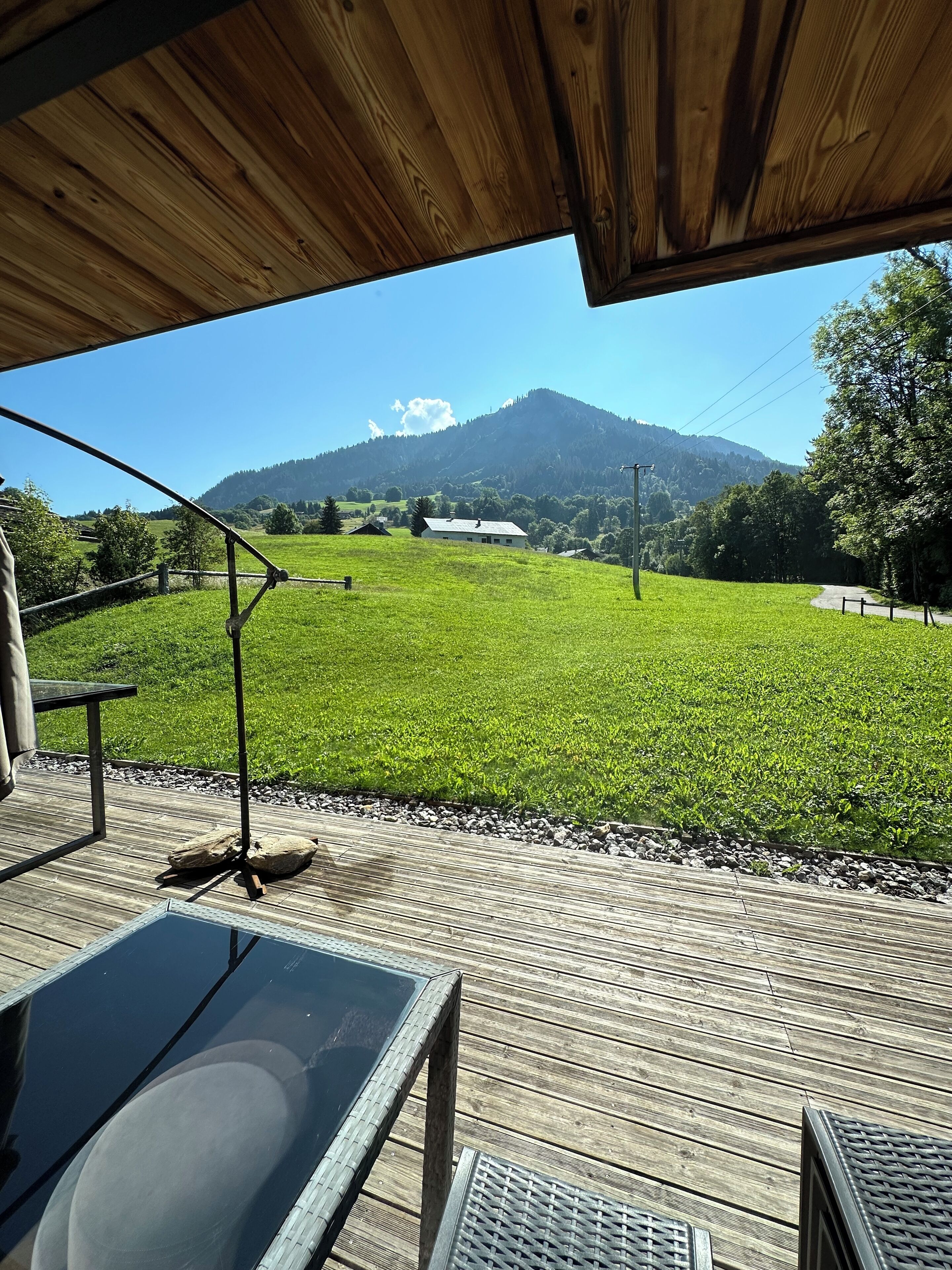 Eine Holzterrasse mit einem Glastisch, mit Blick auf ein grünes Feld und einen Berg.