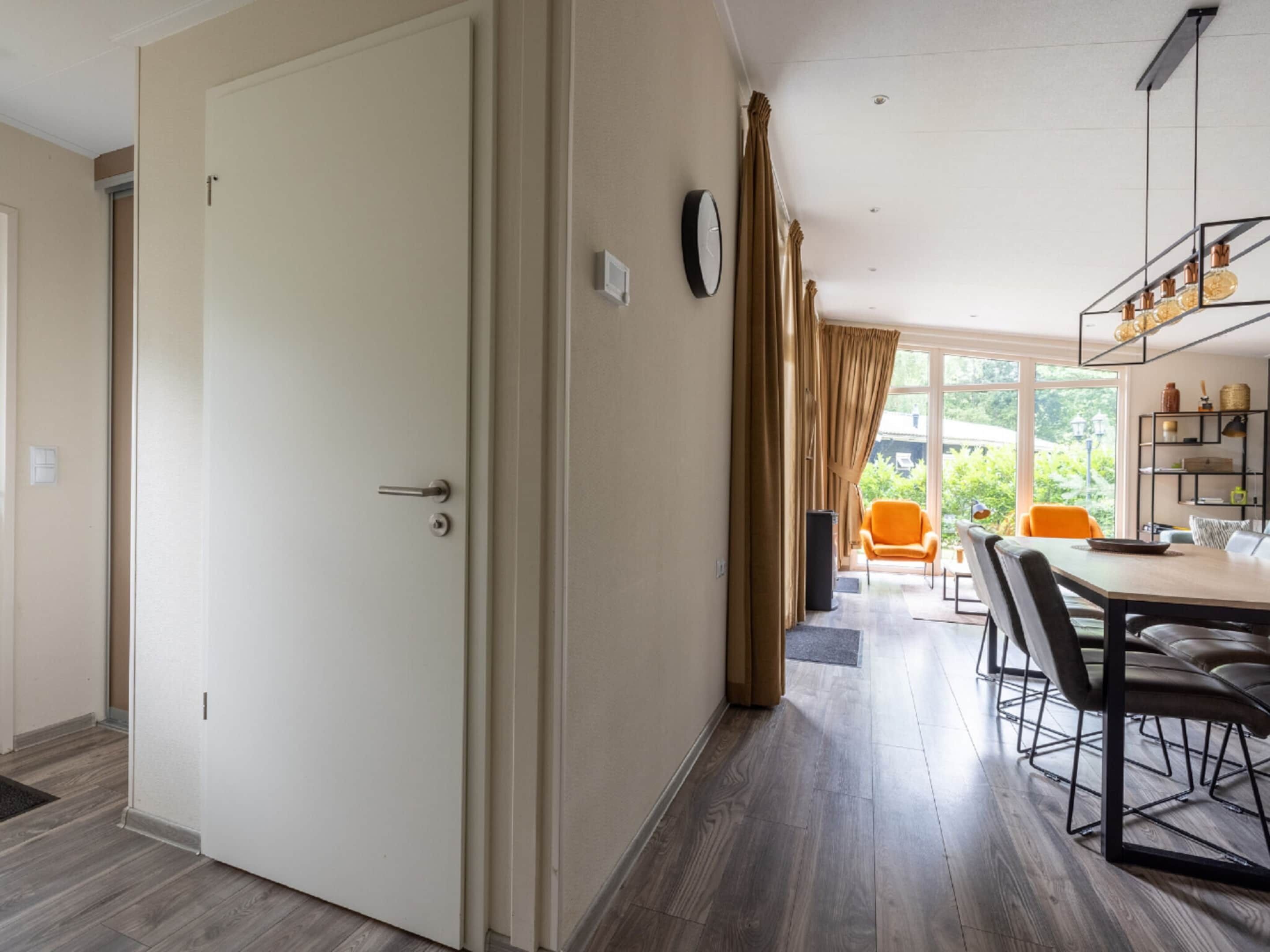 A modern hotel room with a wooden floor, a dining area with a table and chairs, and a view of greenery outside.