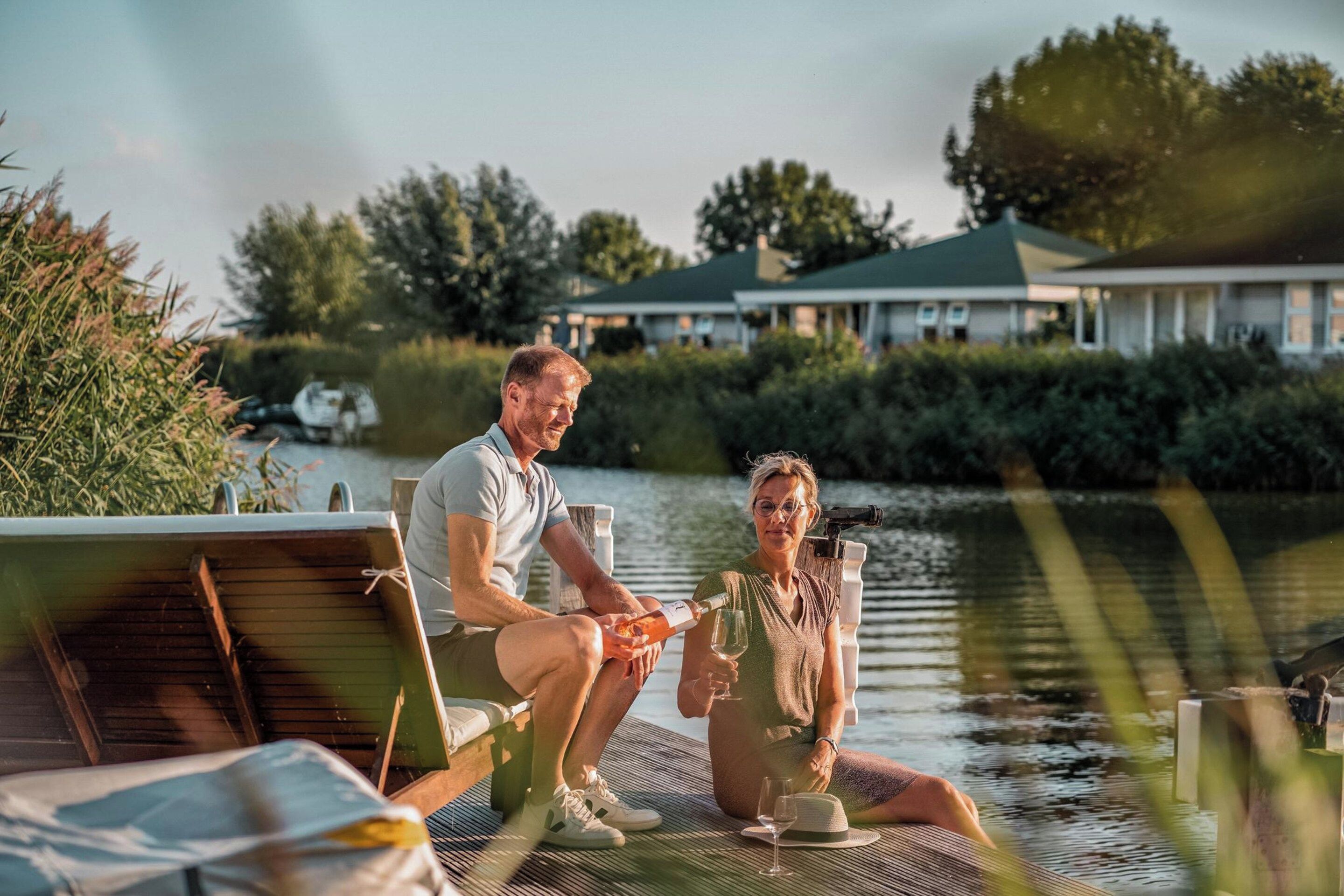 A couple sitting on a dock by a body of water, with a house in the background.