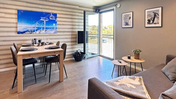 A modern living room with a wooden dining table, a sofa, and a large window with a view of a ski lift.