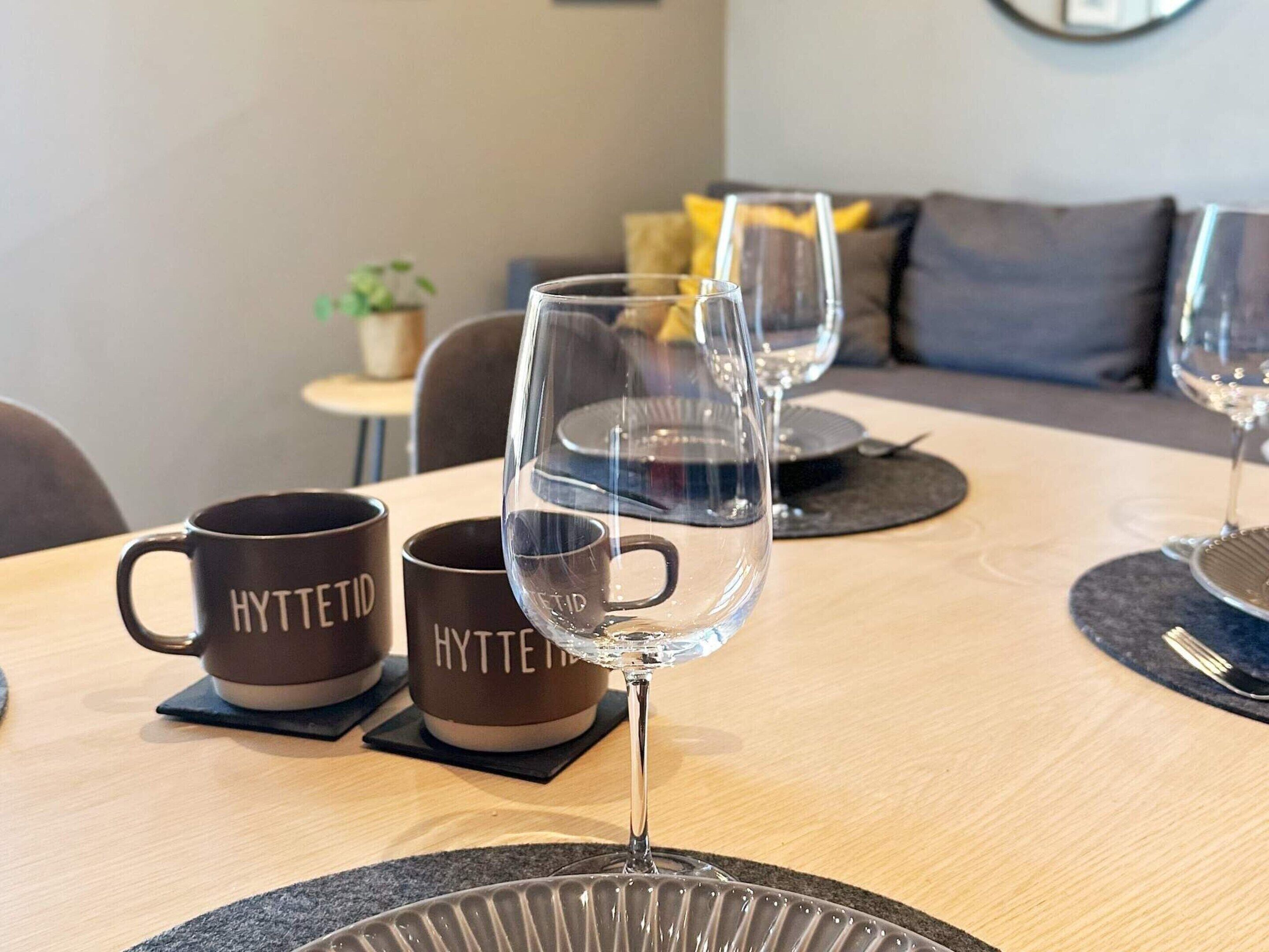 A dining table with three wine glasses and two coffee mugs with the word "HYTTETID" on them.