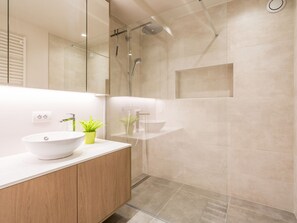 Un baño moderno con un lavamanos blanco, tocador de madera, una planta verde, cabina de ducha de vidrio y un espejo.