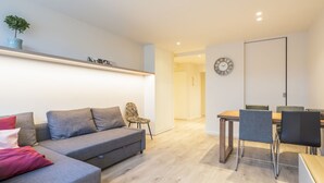 Una sala moderna con un sofá gris, una mesa de comedor con sillas, una planta pequeña en un estante y un reloj en la pared.