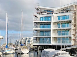 Un puerto deportivo con varias embarcaciones amarradas, un edificio de varios pisos con balcones y un cielo despejado.