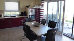 Egy modern konyha, étkezővel, melyben egy nagy ablak, egy piros-fehér csíkos függöny és egy tolóüveg-ajtó található, amely a kültéri teraszra nyílik.