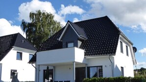 Egy kétszintes ház, sötét színű cserépfedéllel, fedett terasszal, ahol kerti bútorok vannak, és kerítéssel ellátott udvarral.