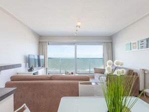 En moderne stue med stort vindue med havudsigt, et fladskærms-tv, en sofa og et spisebord med en vase blomster.