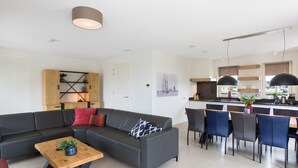 Una sala de estar moderna con un sofá modular, una mesa de centro de madera, y un comedor con sillas azules, con una cocina al fondo.