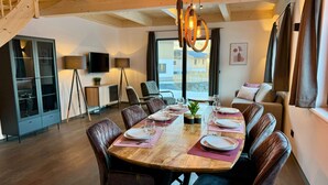 Un coin repas avec une table en bois dressée pour six personnes, entourée de fauteuils en cuir, dans une pièce dotée d’un escalier et d’une grande fenêtre.