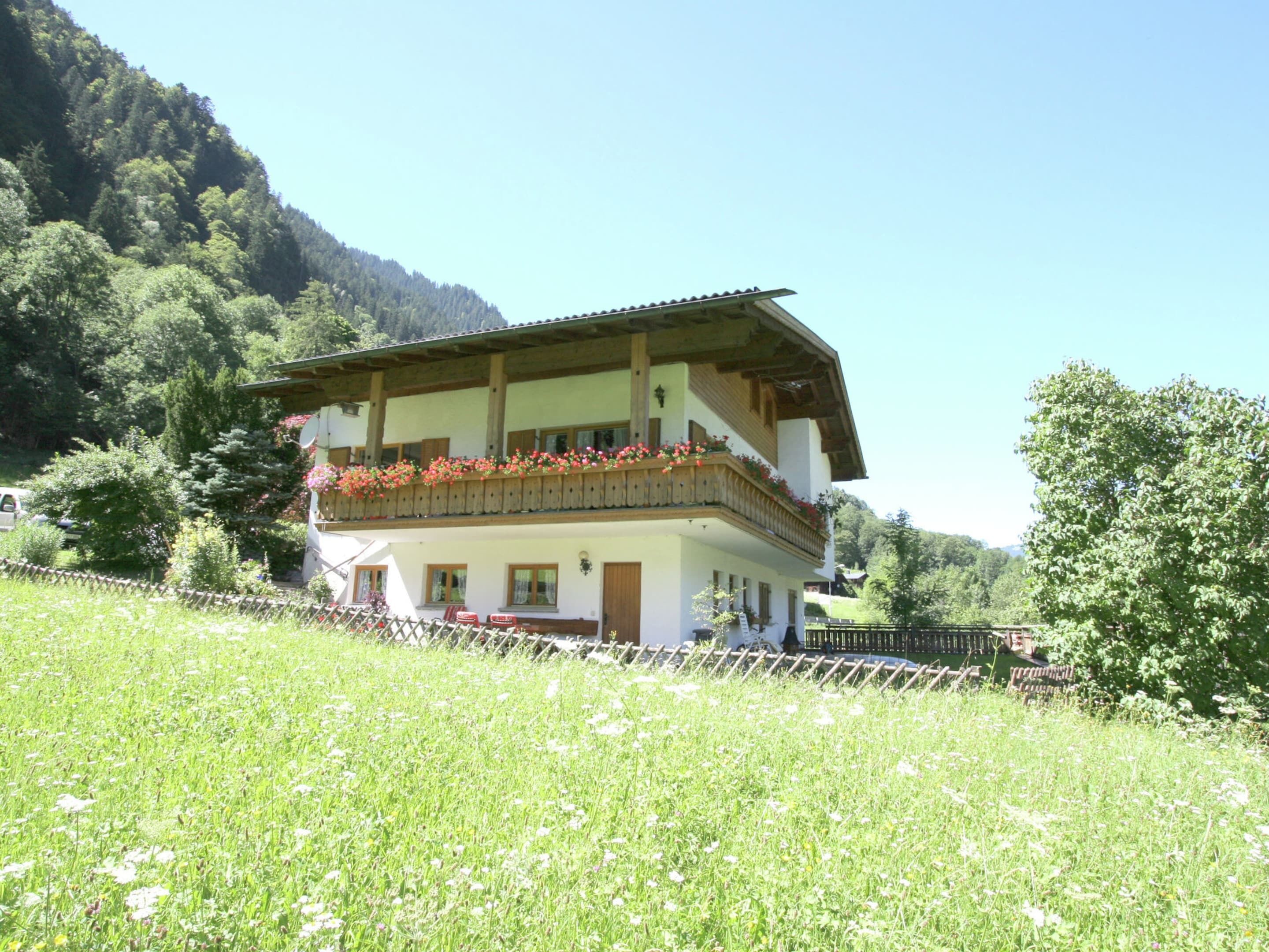 A two-story chalet with a balcony adorned with flowers, surrounded by a lush green landscape and a forested mountain in the background.