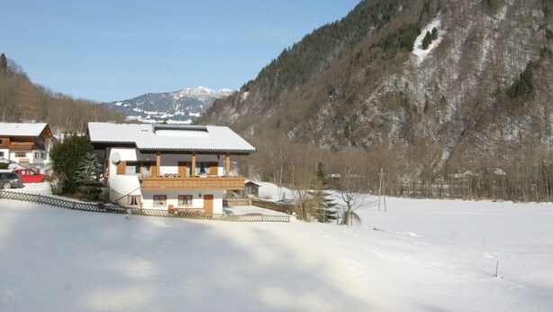 Un edificio de estilo chalé con techo inclinado, rodeado de terreno cubierto de nieve y un paisaje montañoso boscoso.