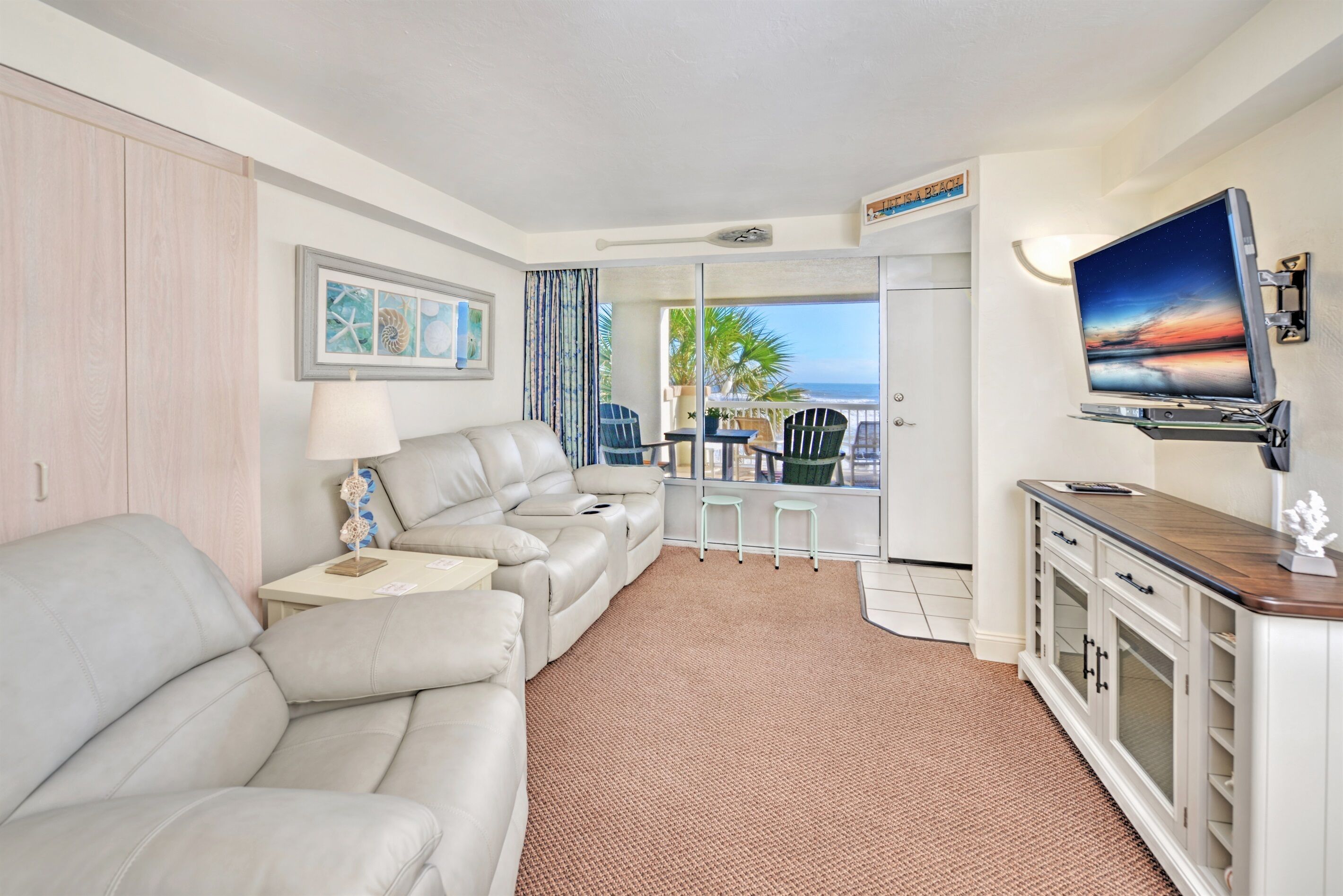 A hotel room with a sofa, a TV, a small table with a lamp, a balcony with a view of the ocean, and a cabinet with drawers.