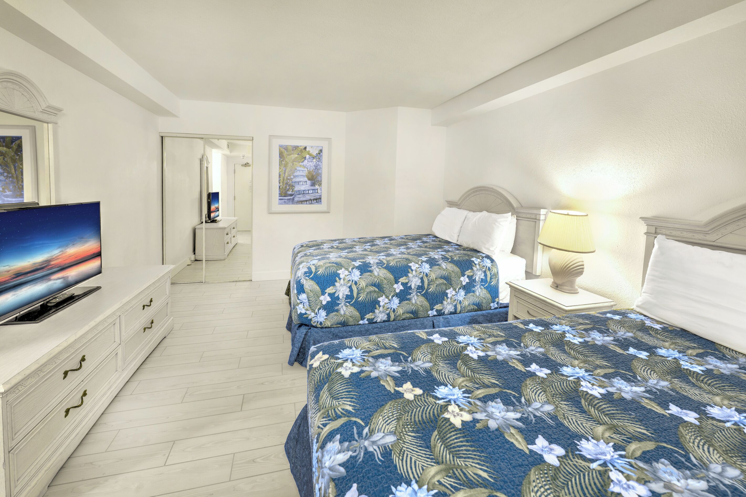 Une chambre d’hôtel avec deux lits, une télévision, une lampe de chevet et des œuvres d’art accrochées aux murs.
