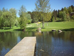 Eine Holzstege, der sich in einen ruhigen Teich erstreckt, umgeben von üppigem Grün und einer Wiese mit Spielgeräten.