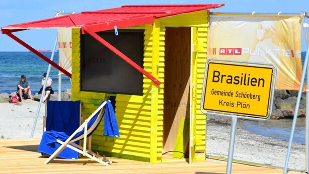 Un chalet de plage avec un toit rouge et des murs jaunes, portant l’inscription « Brasilien Gemeinde Schönberg Kreis Plön ».