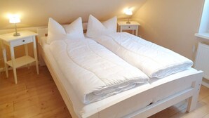 Un lit simple avec une literie blanche, dans une pièce au plancher en bois et aux murs blancs.
