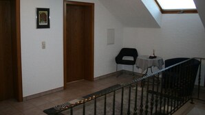 A room with a sloped ceiling, a skylight, a wooden door, a framed picture, a small table with a chair, and a metal railing. - Liefde Voor de Eifel (Huertgenwald)