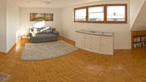 A room with a grey sofa, a white cabinet, a herringbone wooden floor, a painting on the wall, and a window with a view. - Liefde Voor de Eifel (Huertgenwald)